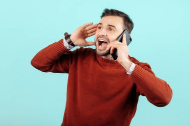 Şok olmuş sakallı genç adam sıradan kıyafetleriyle akıllı telefondan Cyan arka planında konuşuyor..