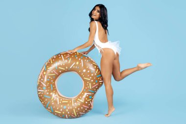 Mayo giymiş çekici genç bir kadın mavi arka planda donut şişirilebilir halkayla mutlu poz veriyor..