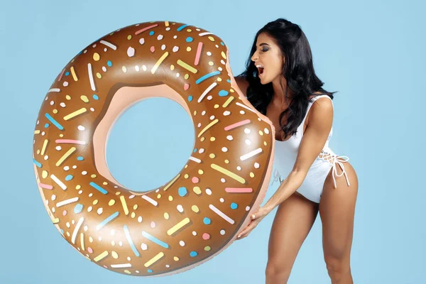 Excited fit young woman dressed in swimsuit posing happy with donut inflatable circle over blue background.