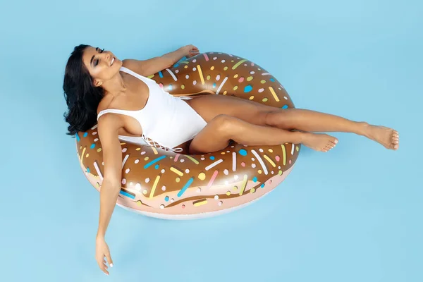 Encantada mujer joven en forma vestida con traje de baño relajante en el círculo inflable de la dona en el fondo azul . — Foto de Stock