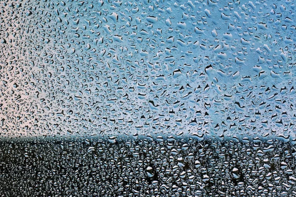 Glass covered with water droplets on a background of blue sky. The lower part is darkened. Soft focus. Rainy season concept.