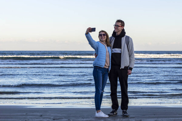 Любящая молодая пара на берегу моря делает селфи с помощью мобильного телефона. Туристы фотографируют во время отпуска в холодное время года
.