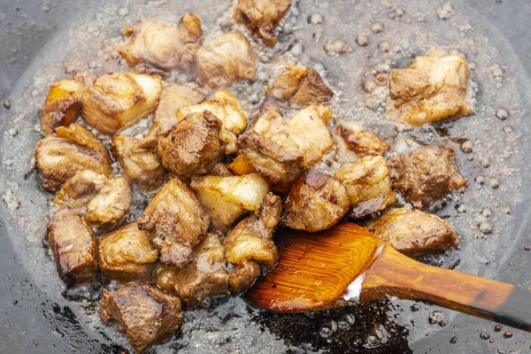 Pieces of red meat are fried in an old frying pan. Close up photo of meat, greaves, wooden kitchen spatula and splashes of fat.