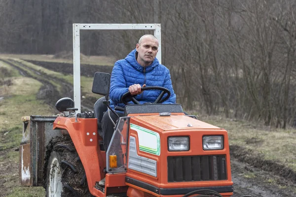 Sad tired tractor driver is sitting on a red tractor without cab during rain in the field near the forest.
