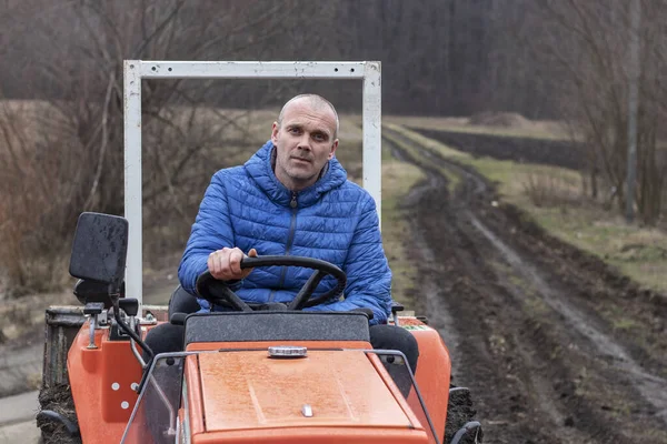 Sad tired tractor driver is sitting on a red tractor without cab during rain. Wet dirt road and winter leafless forest in the background.