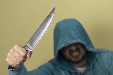 Tıraşsız suratlı, mavi kapüşonlu, elinde büyük bir bıçak tutan kızgın bir adam. Seçici odaklanma, bulanık yüz. Sarı arkaplan.