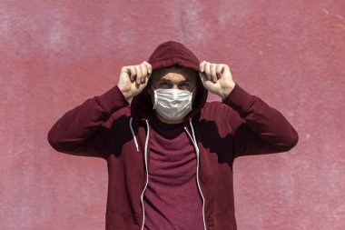 Kırmızı kapüşonlu elbiseli ve yüzünde pembe duvar arkası maskesiyle bir adam. Doğal gün ışığı.