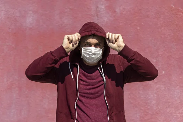 Kırmızı kapüşonlu elbiseli ve yüzünde pembe duvar arkası maskesiyle bir adam. Doğal gün ışığı.