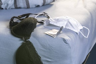 Siyah sutyen, beyaz tıbbi maske ve yatağın kenarında prezervatif ambalajı. Karantina sırasında seks.