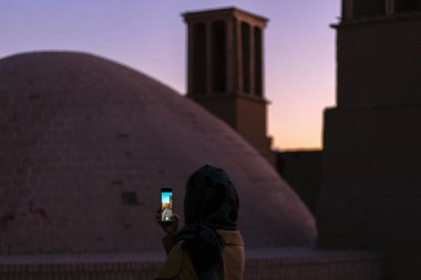 Yazd, İran - Kasım 2019. Kadın turist rüzgar kulesinin (rüzgar yakalayıcı) fotoğraflarını çekiyor - binalarda doğal havalandırma oluşturmak için geleneksel İran mimari unsuru. Gün batımı, karanlık ışık.