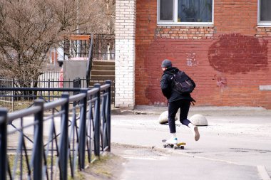 Atletik genç adam. Tuğla bir evin arka planında kaykayla gezen bir hippi. Arkasında bir sırt çantası var.