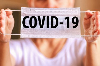 Bir kadın kol mesafesinde tıbbi koruyucu bir maske tutuyor. Maskede COVID-19 yazılı. Bir kadın kendini salgından korumak için ağlıyor. Maskeni takmadan evden çıkma..