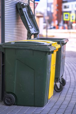 Şehrin sokaklarında duvarın önünde duran plastik çöp konteynırları. Bir konteynerin kapağı açık.