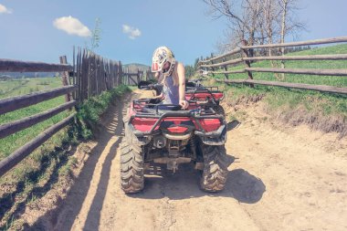 Güneşli bir günde kırsal bir yolda kırmızı bir Atv 'de oturan şapkalı genç bir kadın, kırsal bir yol boyunca tahta bir çit.