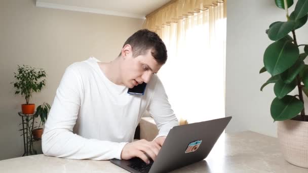 Stresujący młody biznesmen korzystający z laptopa w tym samym czasie rozmawiający przez telefon i czatujący. Rozwiązywanie problemów z pracą, niezadowolony i zirytowany, przysięga na telefon i kładzie telefon na stole — Wideo stockowe