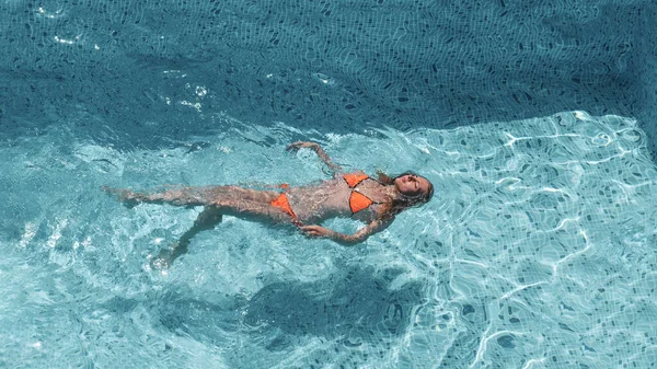 Estate, vacanza, ragazza magra sdraiata sulla schiena in piscina in bikini arancione, vista dall'alto. Acqua cristallina blu — Foto Stock