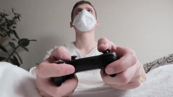 De man met het masker in quarantaine. Koud. Jongeman die thuis op een draadloze controller speelt. Niezen en spelen op. — Stockvideo