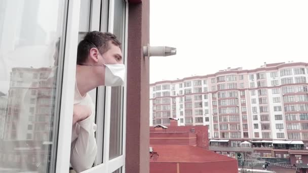 Человек в маске смотрит в окно, глядя на улицу во время болезни. Карантин. Изоляция — стоковое видео