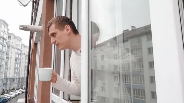 A fiatalember forró kávét vagy teát iszik, miközben kinéz egy többszintes épület ablakán.. — Stock videók