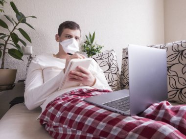 Distance learning. Young Caucasian student in the mask deals for a laptop. Online lecture during quarantine clipart