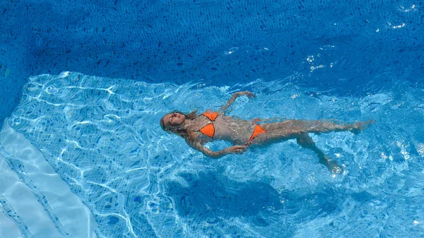 Sommar, semester, smal ung flicka som ligger på rygg i en pool i en orange bikini, ovanifrån. Klarblått vatten — Stockfoto