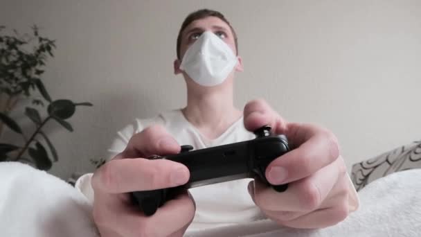 Поняття хвороби та дозвілля. Молодий чоловік у медичній масці на обличчі грає у відеоігри на бездротовому джойстику. Кілька разів, втрачає, засмучується і ставить контролер вниз — стокове відео