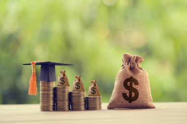 Siyah mezuniyet şapkası, bir masanın üstünde yükselen bozuk paraların üzerinde şapka ve Amerikan Doları çantası. Eğitim fonu, finansal konsept. İleriki çalışmalar için çocuk bilgisi birikimlerini gösterir