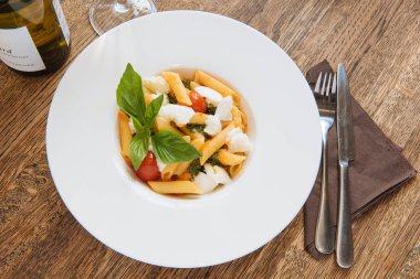 Pappardelle pasta with cheese, tomatoes and basil next to white wine and ingredients on a wooden table in a restaurant clipart