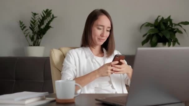 Chica trabajando en casa, escribiendo un teléfono, y sonriendo . — Vídeos de Stock