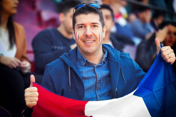 Happy supporter holding french national flag in hands at international sport event - man in the stands of stadium cheering favourite team  soccer fan in arena with blurred people in background