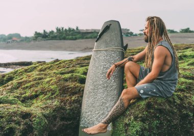 Yanık tenli genç sörfçü dövmeler ve dreadlocks Okyanusu Endonezya, Bali, Canggu, Batu Bolong beach yakın yeşil yosunlu kayalar üzerine oturan ve sörf önce soğutma onun surfboard ile.
