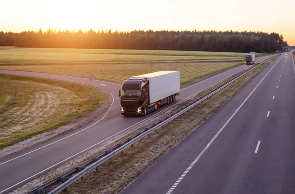 Una caravana de modernos camiones de carga transporta carga por la noche contra el atardecer. Los camiones conducen por la autopista por la noche —  Fotos de Stock