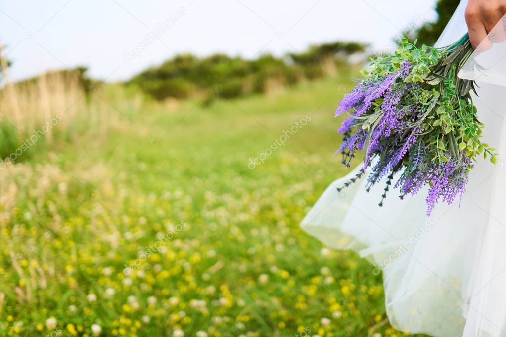 Asian woman wearing white laced bridal dress  holding violet bouquet in front of her at the yellow blossom field located on the cliffs of the beautiful Jeju Island, Korea.                               