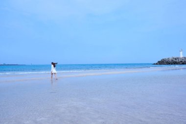 Jeju adasında kayalık platform üzerinde beyaz deniz feneri ile bir plaj yürüyüş Asyalı kadın rüzgarda, onu uzun saçlı beyaz elbiseli.                               