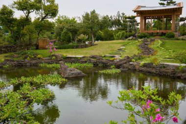  Kayalık adımları, ağaçlar, çim, çiçek, geleneksel gazebo ve Camillia Hill Jeju Adası, Güney Kore çevrede yansıtan bir su birikintisi bahçe                              