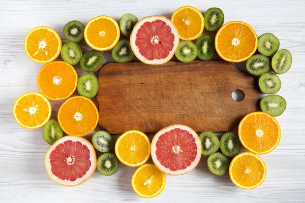 Flat lag. Bovenaanzicht. Houten plank in het midden met gesneden kiwi, sinaasappel, grapefruit en Mandarijn op lichte achtergrond. Zomer achtergrond. — Stockfoto