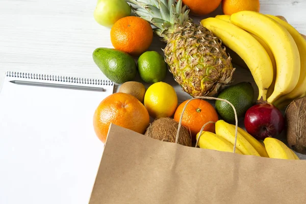 Saco de papel de frutas diferentes com bloco de notas em fundo de madeira branca. Deitado. Vista superior. Espaço de cópia . — Fotografia de Stock