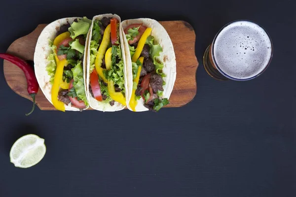 Tacos with fresh vegetables and beer on the board on a black wooden background, top view. Black background for text and design. Flat top view.