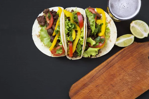 Tacos con carne y verduras, cerveza y lima sobre fondo de madera negra, vista superior. Fondo negro para texto y diseño. Vista superior plana . — Foto de Stock