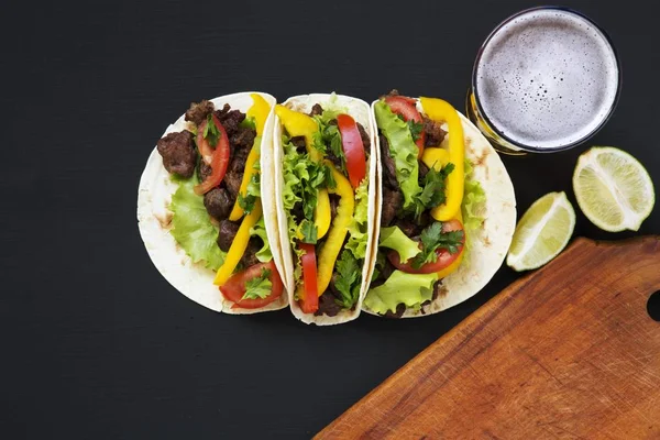 Tacos con carne e verdure, birra e lime su un tavolo di legno nero, vista dall'alto. Sfondo nero per testo e design. Vista dall'alto piatta . — Foto Stock