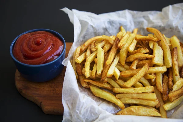 Batatas fritas com ketchup em placa de madeira sobre fundo escuro, vista lateral, close-up — Fotografia de Stock