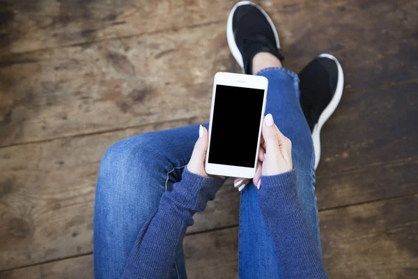 Η κοπέλα κάθεται σε ένα ξύλινο πάτωμα και κατέχει ένα smartphone με μια κενή οθόνη στα χέρια της. Το Top view. — Φωτογραφία Αρχείου