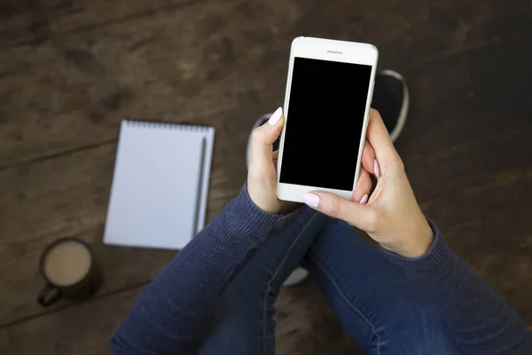 Η κοπέλα κάθεται σε ένα ξύλινο πάτωμα και κατέχει ένα smartphone με μια κενή οθόνη στα χέρια της. Σημειωματάριο και καφέ στο πάτωμα. Το Top view. — Φωτογραφία Αρχείου