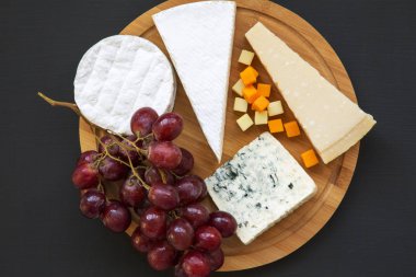 Koyu arka plan üzerinde yuvarlak ahşap tahta üzerinde üzüm ile çeşitli peynirler. Romantik yemek. Düz yatıyordu. Yukarıdan, ana sayfa görünümü.