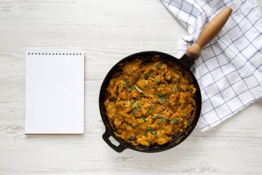 Ev yapımı Tikka Masala tavuğu dökme demir bir tavada, beyaz ahşap arka planda boş bir not defteri, üst manzara. Dümdüz, tepeden, yukarıdan. Boşluğu kopyala.