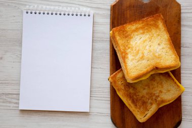 Köy ahşap tahtasında ev yapımı peynirli sandviç, beyaz ahşap yüzeyde boş not defteri, üst manzara. Yukarıdan, dümdüz uzanmış. 
