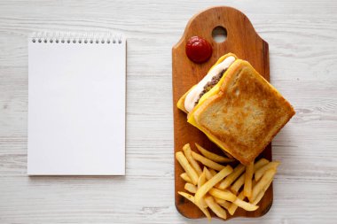 Taze ızgara peynirli burger, yanında patates kızartması, ahşap tahta tahta tahta tahta, beyaz ahşap arka planda boş not defteri, üst manzara. Dümdüz, tepeden, yukarıdan. Boşluğu kopyala.