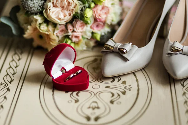 Trouwringen in een mooie rode doos liggen op tafel naast de schoenen van bruid en boeket — Stockfoto