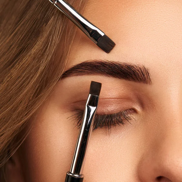 Eyebrows Care. Closeup Of Woman Beautiful Blue Eye, Perfect Shaped Brow, Long Eyelashes With Professional Makeup And Brow Gel Brush. Young Female Model Shaping Brown Eyebrows. High Resolution Image