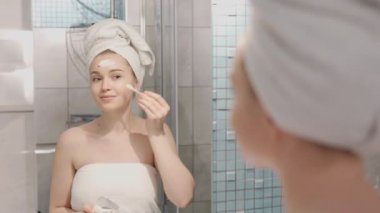 Yüzünde havlu olan mutlu ve çekici bir kadın krem sürdükten sonra makyajını sil ve aynaya bak doğal güzelliğin ve sağlıklı, yumuşak ve yumuşak bir güzelliğin tadını çıkar.
